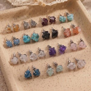 Puede incluir: Una colección de pendientes de botón con piedras preciosas en bruto en varios colores, incluyendo azul, morado, rosa, blanco, negro y naranja. Cada piedra está sujeta en un engaste de metal plateado con garras y poste, dispuestos en filas sobre una bandeja cuadrada.