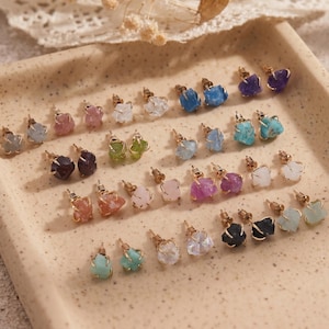 Puede incluir: Una colección de pendientes de botón con piedras preciosas en bruto y sin tallar en varios colores, incluyendo azul, morado, rosa, verde, blanco y negro. Cada piedra está engastada en metal dorado y dispuesta en filas sobre una bandeja beige.