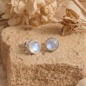May include: A pair of round stud earrings featuring smooth, translucent moonstones set in polished silver metal. Each earring has a circular cabochon stone that displays a soft blue iridescent glow, secured by a simple silver post and backing.