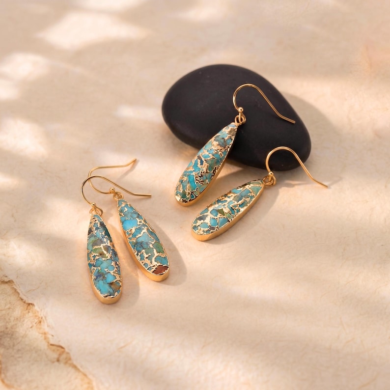 Peut inclure: Boucles d'oreilles pendantes dor&eacute;es avec incrustations de pierres turquoise et dor&eacute;es. Les boucles d'oreilles ont une fermeture &agrave; crochet et sont pr&eacute;sent&eacute;es sur une surface neutre avec une pierre noire en arri&egrave;re-plan. Accessoire de bijouterie.