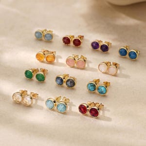 Può includere: Una collezione di orecchini a bottone dorati con varie montature di pietre preziose colorate. Gli orecchini presentano pietre rotonde e lucide nelle tonalità di blu, rosso, viola, verde, arancione, rosa e trasparente. Gli orecchini sono esposti su una superficie neutra.
