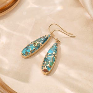 Peut inclure: Une paire de boucles d'oreilles pendantes dor&eacute;es. Chaque boucle d'oreille pr&eacute;sente une pierre turquoise en forme de goutte d'eau avec des veines dor&eacute;es, sertie dans une lunette dor&eacute;e. Les boucles d'oreilles ont une fermeture &agrave; crochet simple.