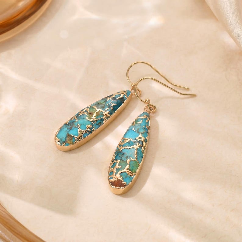 Peut inclure: Une paire de boucles d'oreilles pendantes dor&eacute;es. Chaque boucle d'oreille pr&eacute;sente une pierre turquoise en forme de goutte d'eau avec des veines dor&eacute;es, sertie dans une lunette dor&eacute;e. Les boucles d'oreilles ont une fermeture &agrave; crochet simple.