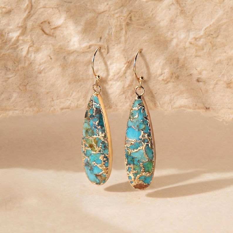 Peut inclure: Une paire de boucles d'oreilles pendantes dor&eacute;es. Chaque boucle d'oreille pr&eacute;sente un incrustation de pierre en forme de goutte d'eau turquoise et dor&eacute;e. Les boucles d'oreilles ont un design simple &agrave; crochet, sur fond neutre.