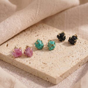 May include: Three pairs of stud earrings featuring raw, unpolished gemstones in pink, turquoise, and black. Each stone is secured by four gold-toned metal prongs. The earrings are arranged in a row, showcasing the natural, irregular texture of the stones.