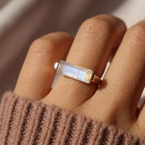 May include: A gold ring featuring a rectangular, iridescent gemstone. The gemstone displays a range of colors, including blue and white. The ring has a simple, elegant design, perfect for everyday wear. The ring is a piece of jewelry.