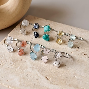 May include: A collection of silver-toned metal ear cuffs, each featuring two raw, uncut gemstones in various colors including clear, dark blue, turquoise, yellow, light blue, orange, and pale pink, arranged in a curved, open-ended design.
