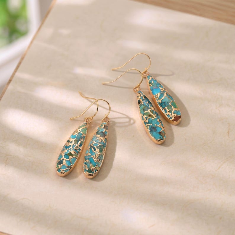 Peut inclure: Deux paires de boucles d'oreilles pendantes dor&eacute;es. Chaque boucle d'oreille pr&eacute;sente une pierre turquoise en forme de goutte d'eau avec des accents dor&eacute;s. Les boucles d'oreilles ont un design simple &agrave; crochet pour oreilles perc&eacute;es.