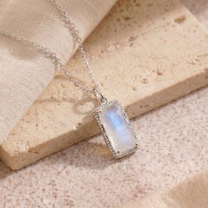 May include: A silver necklace featuring a rectangular pendant with a faceted, iridescent moonstone. The pendant has a beaded metal border and hangs from a delicate silver chain. The stone displays a soft blue glow against the metallic setting.