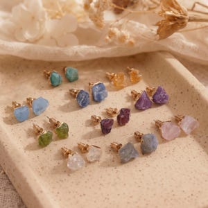 Peut inclure: Une collection de boucles d'oreilles à tige avec des pierres précieuses brutes et non taillées dans diverses couleurs, notamment bleu, vert, violet, jaune et transparent. Chaque pierre est fixée sur un support en métal doré.
