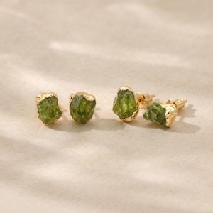 Può includere: Quattro orecchini a bottone placcati in oro con pietre preziose di peridoto verde oliva grezzo. Gli orecchini hanno una forma naturale e organica, con l'incastonatura in oro che accentua la consistenza ruvida delle pietre.