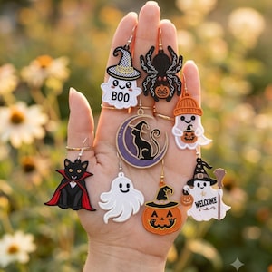May include: A collection of Halloween-themed dangle earrings featuring embroidered designs of ghosts, a black cat in a cape, a spider with a pumpkin, a crescent moon with a cat, a jack-o'-lantern, and a ghost holding a broom with the text WELCOME.