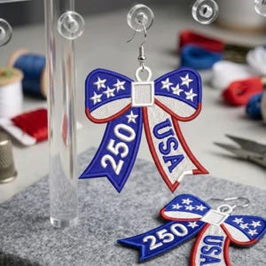 Pode incluir: Brincos bordados em forma de laço nas cores vermelho, branco e azul, com as palavras "USA" e "250". Os brincos têm um tema patriótico com detalhes de estrelas.