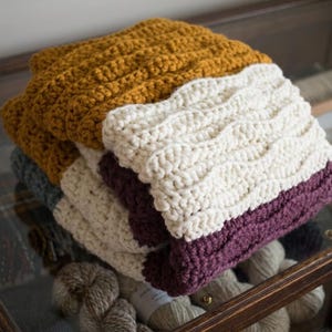 May include: A stack of handmade crochet blankets in various colors. The top blanket is mustard yellow, followed by cream, gray, and deep purple. The blankets have a textured, wavy pattern.