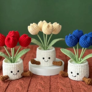 May include: Three crochet tulip flower arrangements in white pots with smiling faces. One has red tulips, one has yellow tulips, and one has blue tulips. The pots have brown arms and are on a brown surface.