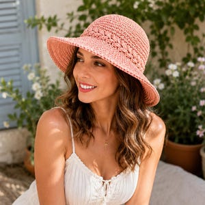May include: A salmon-colored crochet sun hat featuring a braided band detail around the crown. The hat has a rounded top and a wide, downward-sloping brim, crafted from a textured, woven material in a solid, muted coral-pink shade.