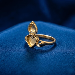 Könnte beinhalten: Ein goldfarbener herzförmiger Medaillonring mit offenem Deckel, der einen leeren Innenraum freilegt. Der Ring hat ein Scharnierdesign, das das Öffnen und Schließen des Deckels ermöglicht. Der Ring ist vor einem dunkelblauen Hintergrund platziert.