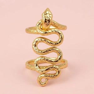 Könnte beinhalten: Ein goldfarbener Ring mit einem Schlangenmotiv, das sich vertikal über zwei Ringbänder windet. Die Schlange hat eine texturierte, geschuppte Oberfläche und einen ausgeprägten Kopf an der Oberseite. Die beiden horizontalen Bänder sind durch den Schlangenkörper verbunden, was eine hohe, geschichtete Struktur mit einer durchgehend polierten, metallischen Oberfläche erzeugt.