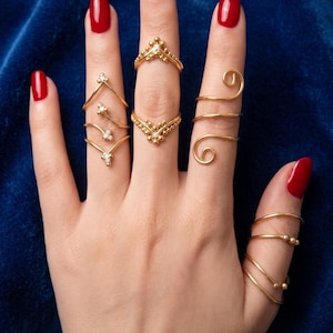Puede incluir: Una mano que muestra varios anillos dorados con diversos diseños. Los anillos incluyen bandas delicadas, formas de chevrón y detalles en espiral. Las uñas están pintadas de un rojo vibrante. Las letras A, B, C y D están junto a los anillos.