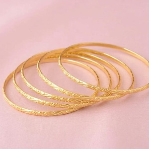Puede incluir: Un conjunto de cinco brazaletes finos de oro, cada uno con un delicado patrón en relieve que se repite a lo largo de toda la superficie exterior. Las pulseras están dispuestas en una formación circular ligeramente superpuesta, mostrando su acabado amarillo metálico.