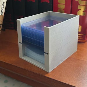 Può includere: Una scatola porta carte grigio chiaro, stampata in 3D, di forma rettangolare. La scatola ha una fessura sul davanti e contiene una pila di bustine per carte bluastre. La scatola è appoggiata su una superficie di legno.