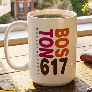 May include: A white coffee mug with the text "BOSTON 617 MASSACHUSETTS" in orange and maroon, filled with coffee. The mug sits on a wooden table next to a bouquet of flowers, a wallet, and keys. The image includes the text "Start your day the Boston way...if you know you know".