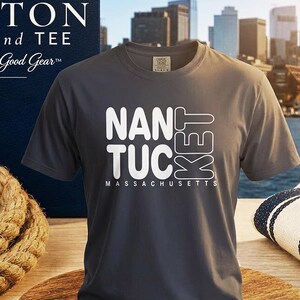 May include: A charcoal gray short-sleeve t-shirt featuring white text that reads NANTUCKET MASSACHUSETTS. The shirt is displayed on a round wooden base. The graphic design includes the word NANTUCKET stacked in a stylized block font.