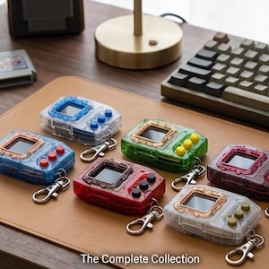 May include: A collection of seven colorful handheld digital pet devices with keychains, arranged on a brown leather desk mat. Each device features a small screen and buttons, with colors including blue, green, red, and clear. The text "The Complete Collection" is at the bottom.
