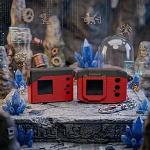 May include: Two red and black electronic device cases with a rectangular screen and buttons. The cases are displayed on a stone surface with blue crystals and small figurines in the background. A silver keychain is attached to one case.