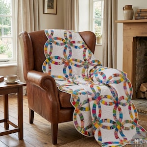 May include: A white quilt with a colorful double wedding ring pattern draped over a brown leather armchair. The quilt features a repeating design of interlocking rings in various colors, including blue, yellow, pink, and green. The chair is in a cozy living room setting.