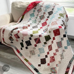 Könnte beinhalten: Eine handgefertigte Steppdecke mit weißem Hintergrund und einem Muster aus bunten Quadraten in Rot, Grün, Blau und Schwarz. Die Steppdecke ist über ein beiges Sofa drapiert und zeigt ihr aufwendiges Design und ihre Nähte. Die Steppdecke hat einen roten Rand.