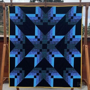 May include: A handmade quilt with a starburst design. The quilt features a black background with geometric star patterns in shades of blue and teal. The edges are finished with a yellow border. The quilt is displayed on a wooden drying rack.