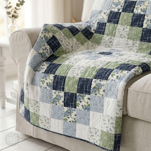 May include: A patchwork quilt draped over a beige sofa. The quilt features a pattern of squares in shades of blue, green, and white. Some squares have floral prints, while others have solid or checkered designs. The quilt adds a touch of comfort and style.