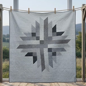 May include: A light gray quilt with a geometric starburst design in shades of gray and black. The quilt is hanging on a clothesline, showcasing its textured surface and stitched details. The quilt's dimensions are not visible.