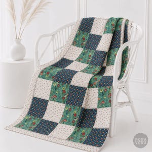 Puede incluir: Una colcha de patchwork hecha a mano sobre una silla de mimbre blanca. La colcha presenta un patrón de tablero de ajedrez con cuadrados de telas florales, azul marino y crema. El borde de la colcha es de color crema con un pequeño estampado floral. La colcha mide aproximadamente 152 cm por 178 cm.