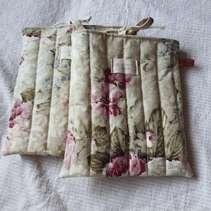 May include: Two rectangular quilted fabric covers featuring a floral pattern with pink, green, and blue flowers on a cream background. Each cover has vertical stitching, a small fabric tag reading cozy cover by emily, and a small red ribbon loop.