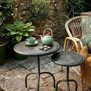 May include: Two round metal side tables with textured surfaces and three curved legs. A light green ceramic teapot with a bamboo handle and a matching teacup on a saucer sit on the larger table, accompanied by four small smooth gray stones.
