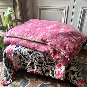 Peut inclure: Une pile de coussins décoratifs et une couverture. Le coussin supérieur est rose vif avec un motif floral et une bordure dorée. La couverture en dessous présente un fond blanc avec des motifs floraux brodés en bleu, rouge et vert.