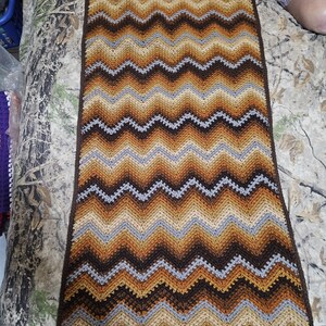 Puede incluir: Una manta de ganchillo hecha a mano con un patr&oacute;n de chevr&oacute;n. La manta presenta un dise&ntilde;o en zigzag repetido en tonos marrones, beige y grises. Los bordes est&aacute;n acabados con un borde festoneado. Esta acogedora manta es perfecta para a&ntilde;adir calidez y estilo a cualquier habitaci&oacute;n.