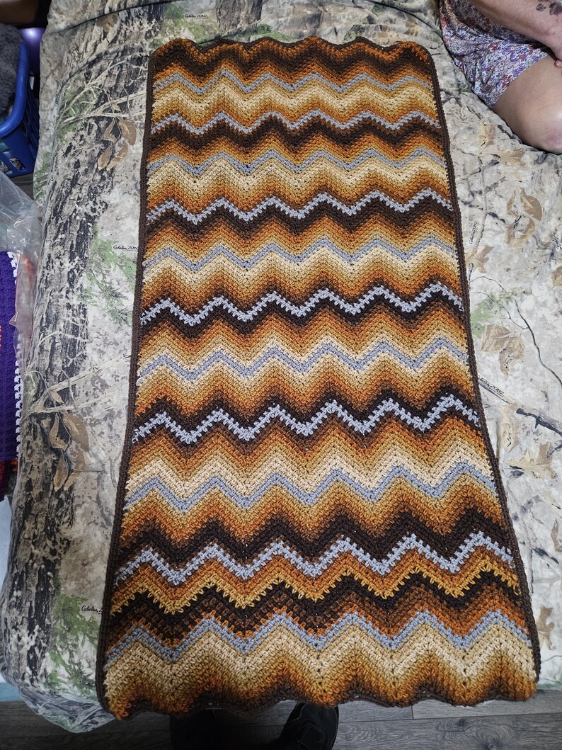 Puede incluir: Una manta de ganchillo hecha a mano con un patr&oacute;n de chevr&oacute;n. La manta presenta un dise&ntilde;o en zigzag repetido en tonos marrones, beige y grises. Los bordes est&aacute;n acabados con un borde festoneado. Esta acogedora manta es perfecta para a&ntilde;adir calidez y estilo a cualquier habitaci&oacute;n.