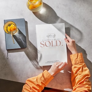 Puede incluir: Una persona sostiene un documento blanco con el texto "SOLD" en una fuente suave y elegante. El documento es una guía para el vendedor. También se ven un cuaderno azul y un vaso de bebida con infusión de naranja.