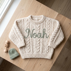 May include: Cream-colored cable knit sweater with the name "Noah" embroidered in green thread. A matching green beanie and a wooden rattle are next to the sweater. The sweater has a classic crew neck and ribbed cuffs.