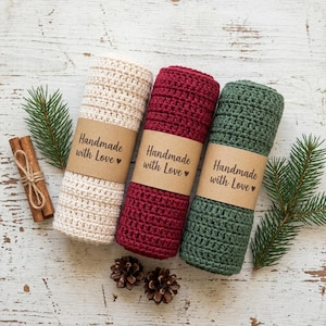 May include: Three rolled crochet washcloths in cream, burgundy, and green, each wrapped with a brown paper band that reads "Handmade with Love." Cinnamon sticks and pine sprigs are arranged on a white, distressed wooden surface.