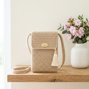 May include: A beige crochet crossbody bag with a flap closure and a long strap. The bag features a gold-colored tassel and a small, gold-colored tag that reads "handmade". The bag is displayed on a wooden shelf next to a white vase with pink flowers.