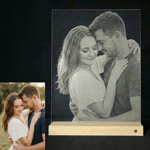Puede incluir: Una foto personalizada grabada en un panel de vidrio transparente, que representa a una pareja abrazándose. La imagen está sobre una base de madera con un tono dorado cálido. Una foto en color más pequeña de la misma pareja es visible en la esquina inferior izquierda.