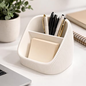 May include: A white desk organizer with a ribbed exterior, holding pens, pencils, and a stack of sticky notes. The organizer has two compartments and is made of a smooth, solid material. A laptop and a potted plant are in the background.