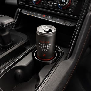 Könnte beinhalten: Eine schwarze Dose "COLD BREW COFFEE BLACK" steht in einem Getränkehalter im Auto. Die Dose hat einen silbernen Deckel und roten und weißen Text. Der Innenraum des Autos ist schwarz mit orangefarbenen Nähten und Klimaanlagenreglern.