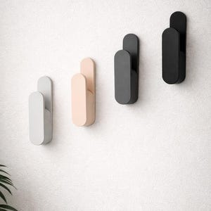 May include: Four contemporary wall hooks in a row, each with a rounded rectangular form. The hooks are in shades of grey, peach, charcoal, and black, mounted on a textured white wall. These hooks are designed for hanging items.