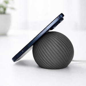 May include: A dark gray, spherical wireless charging stand with a textured, spiraling ribbed surface. A dark blue smartphone is propped against the charger at an angle, with a thin white power cable extending from the base of the charging sphere.