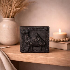 May include: A dark grey, square decorative tile featuring a detailed relief of a camel. The tile has an ornate border and is displayed on a wooden shelf. A lit candle and a vase with dried plants are in the background.
